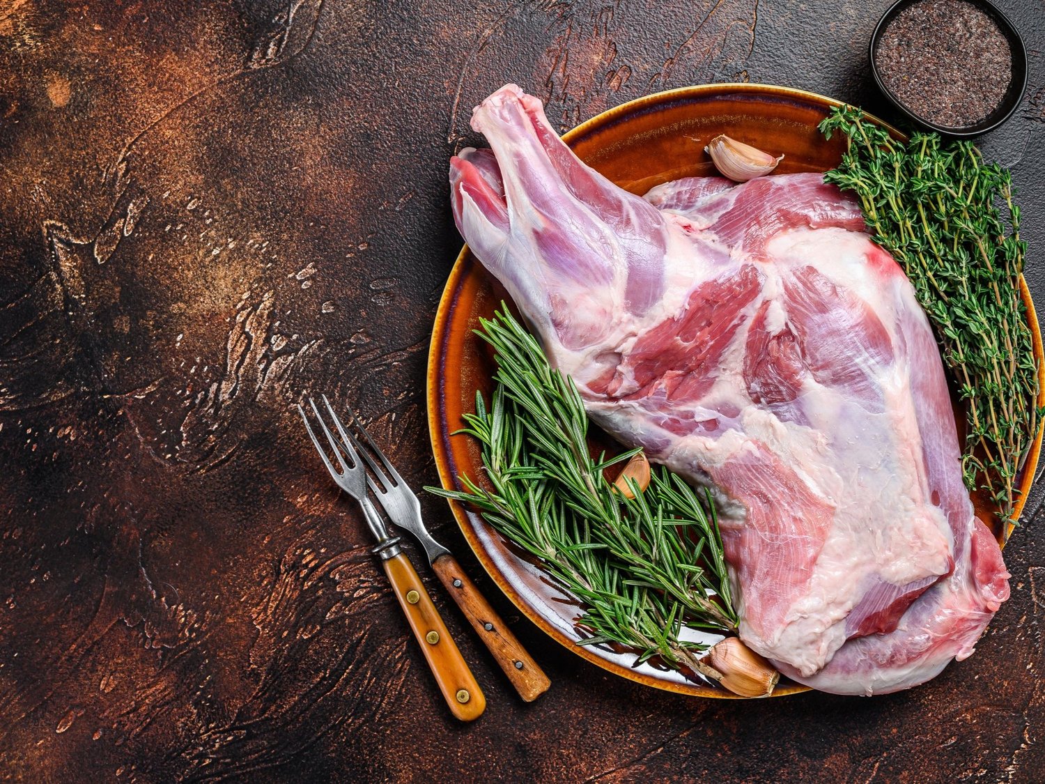 goat_shoulder Raw lamb leg on a rustic plate, garnished with fresh rosemary, thyme, and garlic cloves, accompanied by two forks, with a bowl of black pepper in the background. Perfect for gourmet cooking and meal preparation.