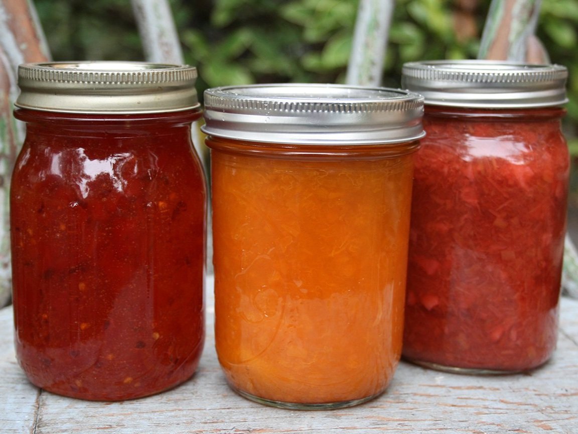 jam_mixed_jars_nolabels1 Three jars of homemade fruit preserves displayed on a rustic wooden surface, featuring a red jam on the left, an orange marmalade in the center, and a red strawberry preserve on the right. The jars are sealed with metal lids, highlighting the vibrant colors and textures of the preserves.