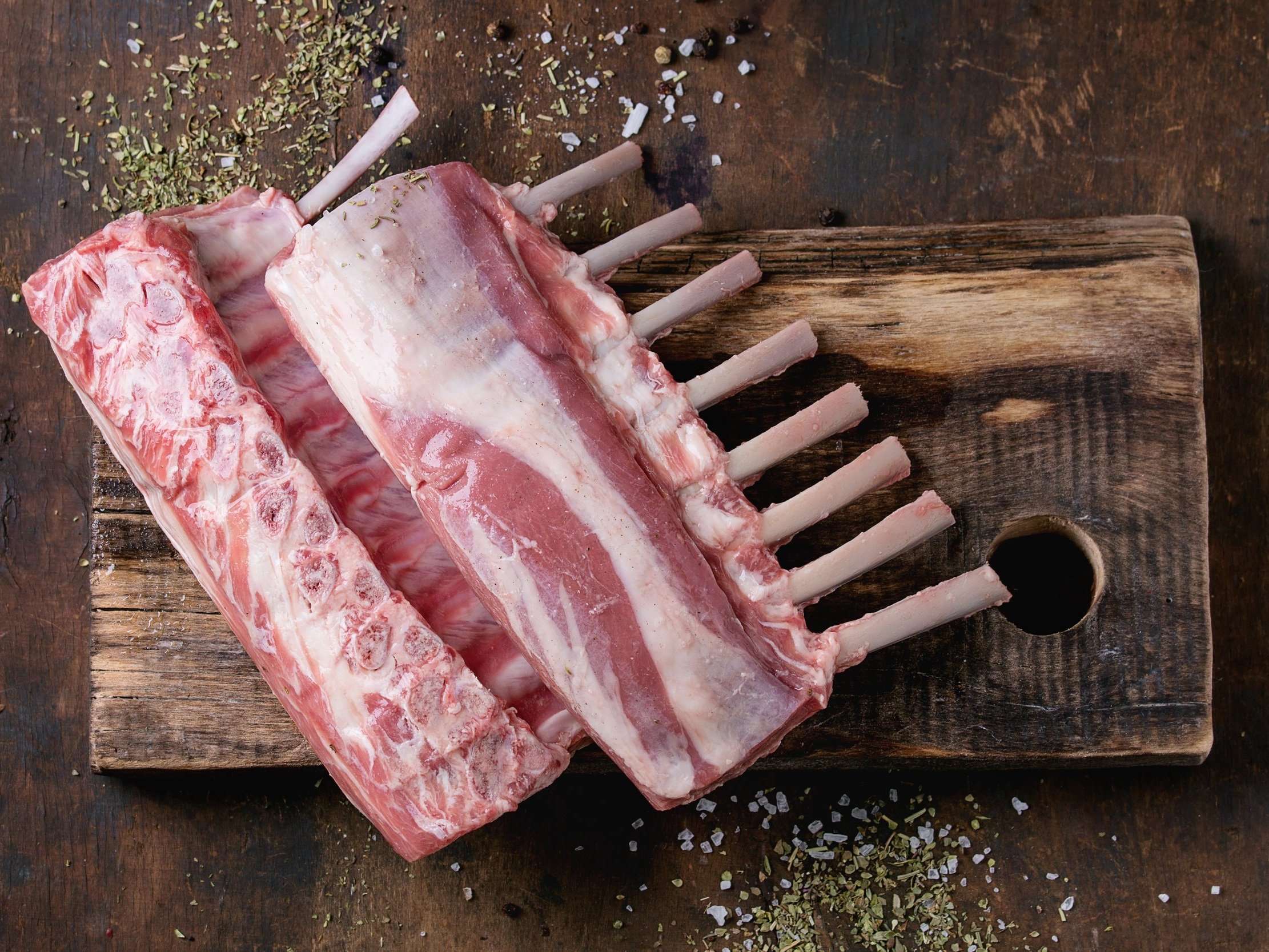lamb_rack_raw Raw rack of lamb resting on a rustic wooden cutting board, garnished with herbs and coarse sea salt, showcasing the meat's marbling and bone structure.