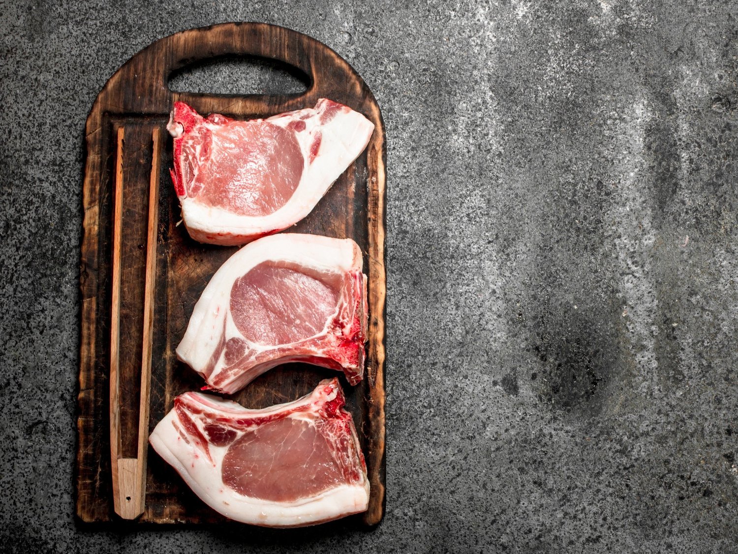 pork_chops_raw_cutting_board Three raw pork chops arranged on a wooden cutting board, with chopsticks placed beside them, set against a textured gray background.